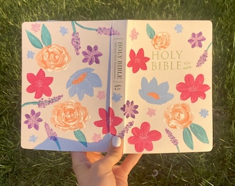 May include: A white book with a floral design on the cover. The book is titled "HOLY BIBLE" with "Old and New Testaments" and "KJV" printed on the spine. The book is open to a page with a floral design.