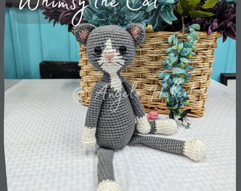 Puede incluir: Un gato de crochet gris con detalles blancos se sienta sobre una superficie con un patrón blanco y gris. El gato tiene ojos negros y una nariz rosa. El gato está sentado junto a una cesta de mimbre llena de flores verdes y moradas.