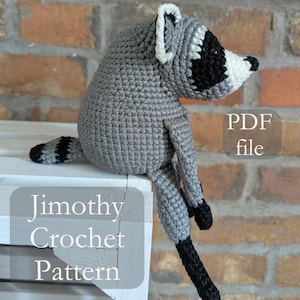 May include: A gray and white crocheted raccoon toy with black eye patches and a striped tail. The text on the image reads PDF file and Jimothy Crochet Pattern. The raccoon has long limbs and is positioned in a seated posture with its tail extended.