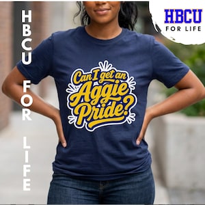 May include: A navy blue short-sleeved t-shirt featuring a yellow and white graphic print on the chest. The text reads, Can I get an Aggie Pride? in a stylized script font, surrounded by white decorative line accents and a circular border.