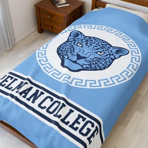 May include: A light blue blanket featuring a jaguar head design within a white circular border. The words "ELMAN COLLEGE" are printed in navy blue on a white stripe at the bottom of the blanket. The blanket is on a bed.