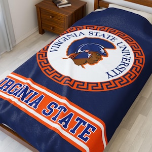 May include: A navy blue blanket featuring the Virginia State University logo. The logo is a circular design with the words "VIRGINIA STATE UNIVERSITY" in a white font, and a graphic of a Trojan warrior. The blanket also has the words "VIRGINIA STATE" in a white font.