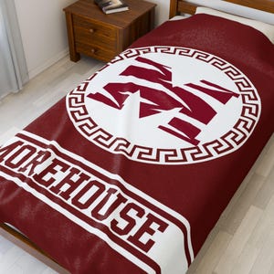 May include: A maroon fleece blanket with a white circle containing a maroon "M" and a white Greek key border. The words "Morehouse" are printed in white on the bottom of the blanket.