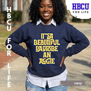 May include: A navy blue long-sleeve sweatshirt featuring a white collared shirt underneath. The sweatshirt displays yellow text that reads IT'S A BEAUTIFUL DAY TO BE AN AGGIE. The design is centred on the chest with a bold, stylised font.