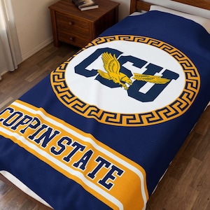 May include: A navy blue blanket with a large circular emblem featuring the letters "CSU" and a yellow eagle. The emblem is surrounded by a gold Greek key design. The words "COPPIN STATE" are displayed in white and gold.