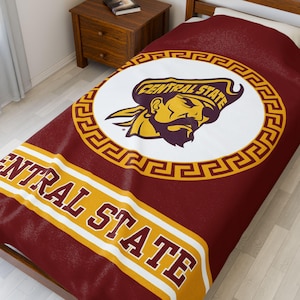 May include: A maroon and gold fleece blanket with a white circle featuring a pirate mascot and the text "Central State". The circle is surrounded by a gold Greek key pattern. The bottom of the blanket has the text "Central State" in white and gold.