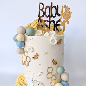 Può includere: Una torta bianca decorata con un personaggio di Winnie the Pooh, favi, api e margherite bianche. La torta è sormontata da un topper dorato con glitter che dice "Baby Asher".