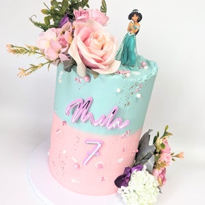 Pode incluir: Um bolo de dois andares decorado com glacê rosa e azul. O andar superior é azul claro com um arranjo floral rosa e uma figura da princesa Jasmine do filme de animação Aladdin da Disney. O andar inferior é rosa com um número 7 rosa neon e o nome "Mila" escrito em letras neon rosas. O bolo está sobre um tabuleiro de bolo branco.