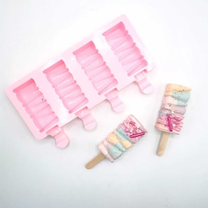 Może przedstawiać: Różowa silikonowa forma na lody na patyku z czterema wnękami i drewnianymi patyczkami. Obok formy pokazane są dwa kolorowe lody na patyku z posypką i wirami.