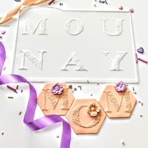 Könnte beinhalten: Eine transparente Acrylform mit dem Wort "MONDAY" in einem geprägten, spitzenartigen Schriftzug. Darunter sind drei pfirsichfarbene, sechseckige Kekse mit floralen Akzenten und den Buchstaben "M", "C" und "M" verziert. Ein lila Band und Streusel vervollständigen die Szene.