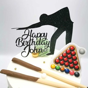Może przedstawiać: Biały tort ozdobiony czarną sylwetką osoby grającej w bilard, stołem bilardowym z czerwonymi kulami i dwoma patyczkami pokrytymi czekoladą. Tort jest ozdobiony zieloną i złotą dekoracją oraz napisem "Happy Birthday John".