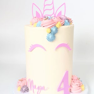 Puede incluir: Un pastel blanco decorado con glaseado rosa, azul y amarillo. El pastel tiene un cuerno y orejas de unicornio rosas en la parte superior, y el número 4 y el nombre "Maya" escritos en glaseado rosa.