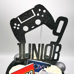 May include: Black and white cake topper with a video game controller and the word "JUNIOR" in glitter. The cake is decorated with a Lego logo, a PlayStation logo, and a white PlayStation controller.