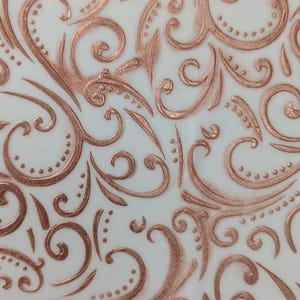 May include: Close-up of a white surface with a repeating pattern of raised, copper-coloured swirls and dots. The design is reminiscent of ornate, vintage decor, with a textured, three-dimensional effect.