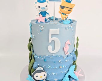 May include: A two-tiered blue birthday cake decorated with a sea-themed design. The cake features a white number "5" on the top tier, and is topped with figurines of characters from the cartoon "The Octonauts". The cake is decorated with blue and white frosting, sprinkles, and sea-themed decorations.