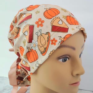 Puede incluir: Un gorro quirúrgico beige con un estampado repetido de calabazas naranjas, rebanadas de pastel, flores y un pavo asado. El gorro tiene un diseño de lazo y se usa en una cabeza de maniquí.