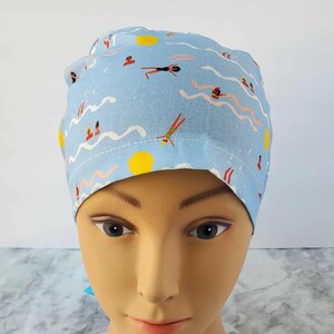 May include: A light blue scrub cap featuring a whimsical pattern of swimmers, suns, and waves. The design includes figures in red, black, and green, with yellow suns and white wave lines. This cap is designed for medical professionals.
