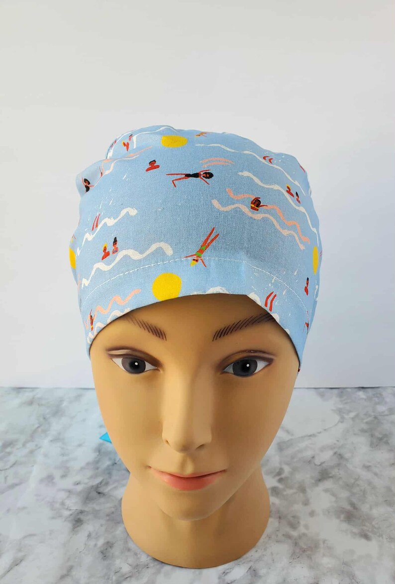 May include: A light blue scrub cap featuring a whimsical pattern of swimmers, suns, and waves. The design includes figures in red, black, and green, with yellow suns and white wave lines. This cap is designed for medical professionals.