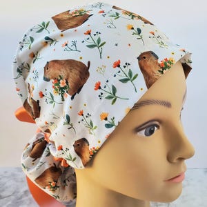 Puede incluir: Gorro quirúrgico blanco con un estampado repetido de marmotas marrones sosteniendo ramos de flores, con detalles florales naranjas y amarillos. El gorro tiene un lazo de cinta naranja.