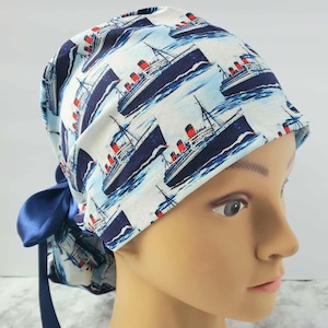 May include: A white and blue patterned scrub cap featuring a repeating design of navy blue cruise ships with red smokestacks. The cap has a navy blue satin bow at the back. The cap is worn on a mannequin head.