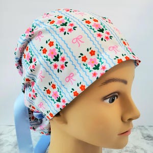 Puede incluir: Un gorro quirúrgico blanco con un estampado floral y de lazo en rosa, rojo y verde, acentuado por rayas onduladas de color azul claro. El gorro tiene una cinta azul claro.