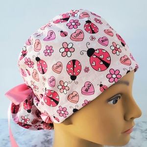 May include: Pink surgical scrub hat with a pink and white print of ladybugs, hearts, and flowers. The text "LOVE" and "BUG" are repeated throughout the print.