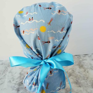 May include: A light blue scrub cap with a repeating pattern of swimmers, waves, and yellow suns. The cap is tied with a bright blue satin ribbon. The fabric has a vintage, slightly faded look.