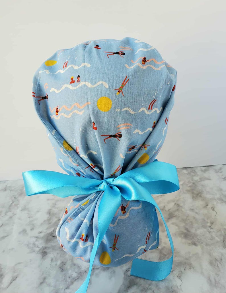 May include: A light blue scrub cap with a repeating pattern of swimmers, waves, and yellow suns. The cap is tied with a bright blue satin ribbon. The fabric has a vintage, slightly faded look.
