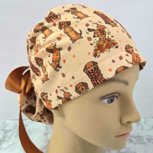 Puede incluir: Un gorro quirúrgico de color beige con un patrón repetitivo de perros salchicha marrones que llevan jerséis y bufandas, rodeados de hojas de otoño, estrellas y corazones pequeños. El gorro incluye una cinta marrón lisa en la parte trasera.