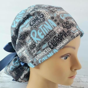 Puede incluir: Un gorro de quirófano gris y azul con estampado y un lazo azul. El gorro presenta una variedad de términos médicos relacionados con los ojos, incluyendo "retina", "córnea", "cataratas", "láseres" y "oftalmología".