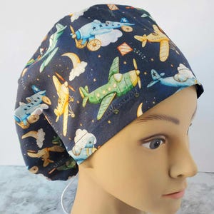 May include: Navy blue surgical scrub cap with a whimsical airplane pattern. The cap features various colorful airplanes, clouds, stars, and moons. The airplanes are in shades of blue, green, and yellow. The cap is designed to be worn in a medical setting.