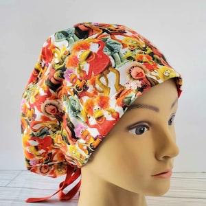 May include: A red, white, and yellow surgical scrub hat with a colorful pattern of cartoon characters. The hat has red ties.