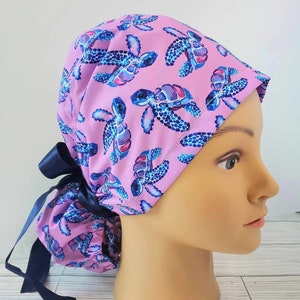May include: A pink surgical scrub hat with a blue and purple sea turtle pattern. The hat has a navy blue ribbon tie.