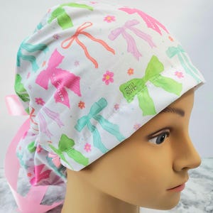 Op de afbeelding: Een witte scrub cap met een patroon van kleurrijke strikken in roze, groen en teal, samen met kleine roze bloemen. De cap heeft een roze lint en de tekst "Me is Sisen" is zichtbaar aan de zijkant.