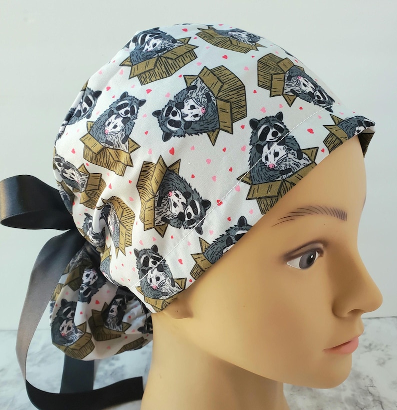 May include: A white scrub cap featuring a repeating pattern of raccoons in cardboard boxes and small pink hearts. The cap has a black satin ribbon tie in the back. The raccoons are gray and white with black masks.