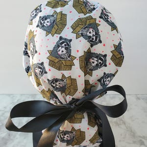 May include: A white scrub cap featuring a repeating pattern of raccoons in cardboard boxes and small pink hearts. The cap is tied with a large black satin bow.