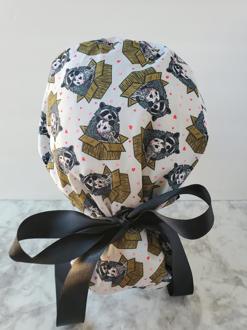 May include: A white scrub cap featuring a repeating pattern of raccoons in cardboard boxes and small pink hearts. The cap is tied with a large black satin bow.