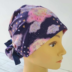 Puede incluir: Un gorro de quirófano morado con un lazo en la parte posterior. El gorro tiene un patrón de nubes rosa y blancas con purpurina dorada y corazones rosas.