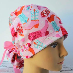 May include: Pink surgical scrub hat with a pattern of colorful cowboy boots and white flowers. The hat has a tie at the back.