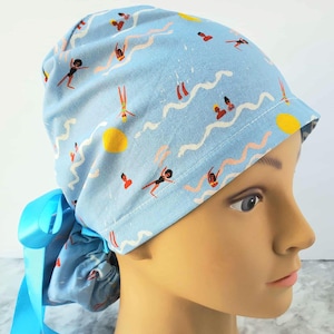 May include: Light blue scrub cap with a repeating pattern of swimmers, suns, and wavy lines. The cap is tied with a light blue ribbon. The design features figures in black swimsuits and orange swimsuits. The cap is designed to be worn by medical professionals.