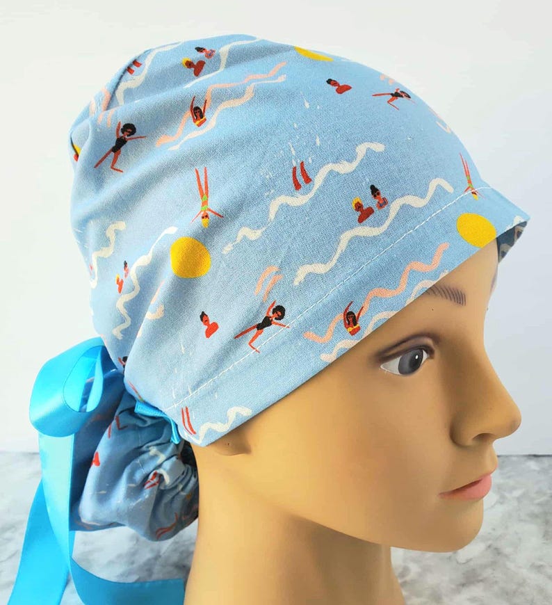 May include: Light blue scrub cap with a repeating pattern of swimmers, suns, and wavy lines. The cap is tied with a light blue ribbon. The design features figures in black swimsuits and orange swimsuits. The cap is designed to be worn by medical professionals.