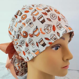 Puede incluir: Un gorro de quirófano blanco con un patrón de tazas de café naranjas, donuts y hojas de otoño. El gorro tiene un lazo de satén naranja a juego.