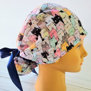 Puede incluir: Un gorro de quir&oacute;fano blanco con un estampado colorido de gatos de dibujos animados. El gorro tiene una cinta azul para atar.