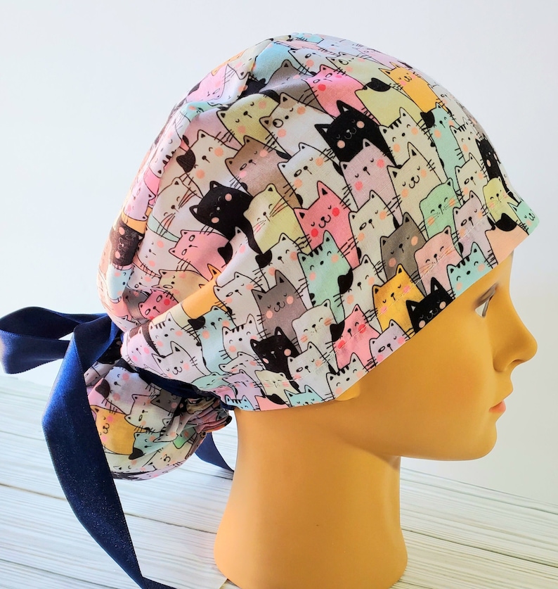 Puede incluir: Un gorro de quir&oacute;fano blanco con un estampado colorido de gatos de dibujos animados. El gorro tiene una cinta azul para atar.