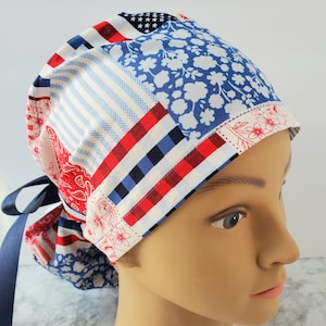 May include: A patchwork scrub cap with a patriotic theme. The cap features a mix of red, white, and blue patterns, including stripes, floral prints, and stars. A navy blue ribbon is attached for tying. This cap is designed for medical professionals.