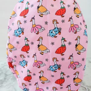 May include: Pink fabric item patterned with cartoon geese wearing various outfits, including dresses and hats. The geese are in orange, pink, blue, and red, with small flower accents. The background is a solid pink.