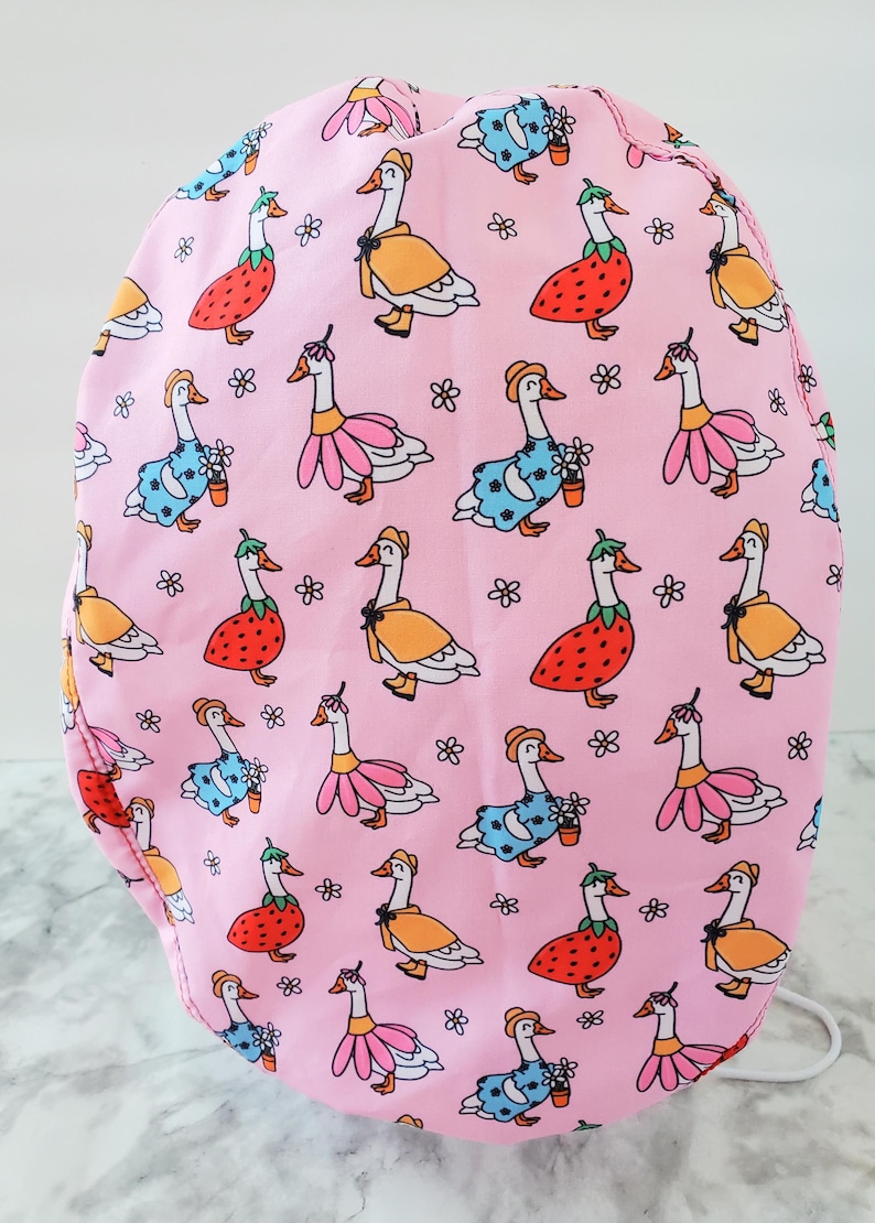 May include: Pink fabric item patterned with cartoon geese wearing various outfits, including dresses and hats. The geese are in orange, pink, blue, and red, with small flower accents. The background is a solid pink.