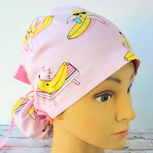 以下が含まれることがあります： ピンクの外科用スクラブハットに、楽しいバナナのプリントが施されています。バナナはサングラスをかけて、ビーチチェアでリラックスしています。