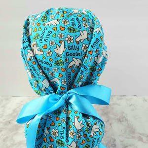 May include: A light blue scrub cap with a pattern of white geese, flowers, and hearts. The words "Silly Goose!" are printed on the cap. A light blue ribbon is tied in a bow at the back.