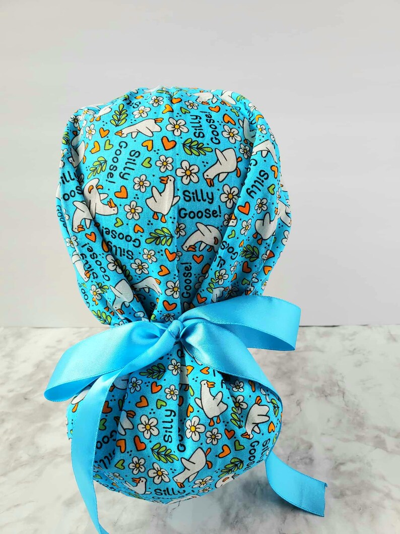 May include: A light blue scrub cap with a pattern of white geese, flowers, and hearts. The words "Silly Goose!" are printed on the cap. A light blue ribbon is tied in a bow at the back.